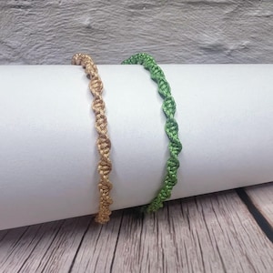 May include: Two handmade macrame bracelets. One is a light brown color, and the other is green. The bracelets are displayed on a white cylindrical surface against a textured gray background. The bracelets are made of knotted cord.