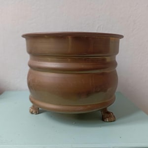May include: A bronze-coloured planter with a rounded body and a wide rim. The planter rests on three small, claw-shaped feet. The surface has a slightly aged appearance, suggesting it may be vintage or antique. This decorative piece is suitable for indoor or outdoor use.