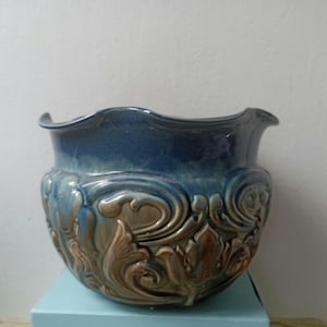 May include: A ceramic planter featuring a deep blue glazed upper section and a lower body adorned with raised, intricate tan and brown scrollwork patterns. The vessel has a wide, scalloped rim and a rounded shape, showcasing a glossy finish.