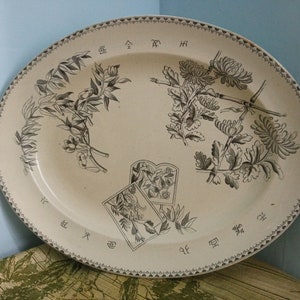 May include: An oval ceramic platter with a cream-coloured background and black floral designs. The platter has a decorative border with a repeating pattern of small triangles. The floral designs include chrysanthemums, bamboo, and other plants. The platter has Japanese characters around the edge.