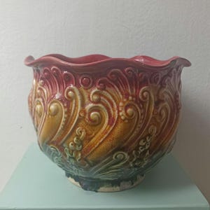 May include: A decorative ceramic planter with a ruffled rim. The planter features a detailed embossed design with a colour gradient of red, yellow, and green. The base is a light blue colour.