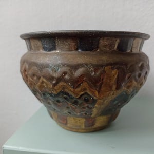 May include: A handcrafted ceramic pot with a wide, rounded form. The pot has a textured surface with brown, tan, and blue hues. Decorative patterns and relief designs adorn the exterior, adding visual interest to this unique piece.