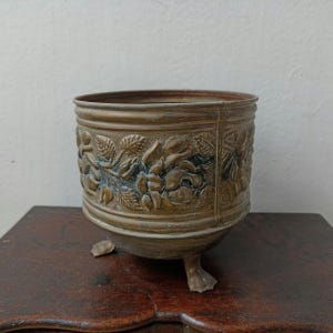 May include: A vintage bronze-coloured planter with a floral relief design. The planter has a cylindrical shape, three small feet, and a detailed pattern of roses and leaves around the exterior. The aged metal has dark accents.
