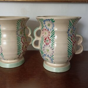 May include: Two cream-coloured ceramic vases with floral designs. The vases have two handles on each side and a green base.