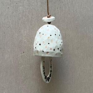 May include: White ceramic bell with black and pink polka dots and a gold accent. The bell is hanging from a brown cord.