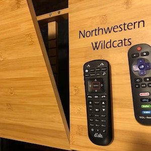 May include: Two black remote controls on a wooden surface. The remote on the left has the word "dish." printed on it. The remote on the right has buttons for Netflix, Sling, Hulu, Vudu, and TCL Roku TV. The surface has the words "Northwestern Wildcats" printed on it.