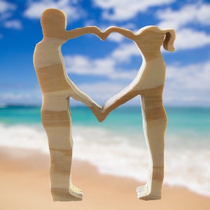 Puede incluir: Dos figuras de madera se encuentran de frente, formando un corazón con sus brazos. Las figuras se recortan sobre un fondo borroso de una playa con agua azul y nubes blancas.