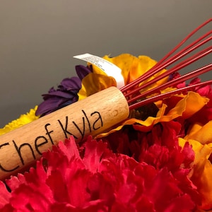 May include: A wooden rolling pin with the text "Chef Kyla" engraved on it. The rolling pin is surrounded by red, yellow, and purple artificial flowers.