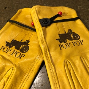 May include: A pair of yellow leather work gloves with a black tractor design and the text "POP POP" on each glove.