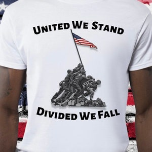 May include: White t-shirt featuring the text "UNITED WE STAND DIVIDED WE FALL" in black. The shirt displays a graphic of soldiers raising the American flag. The flag is red, white, and blue.