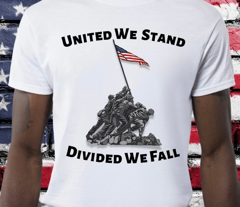 May include: White t-shirt featuring the text "UNITED WE STAND DIVIDED WE FALL" in black. The shirt displays a graphic of soldiers raising the American flag. The flag is red, white, and blue.