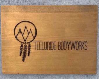 May include: A wooden sign with a brown background and a black design. The design features a circle with three mountain peaks inside, and three feathers hanging from the bottom. The text "TELLURIDE BODYWORKS" is printed below the design.