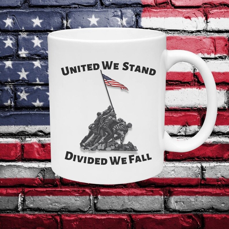 May include: White ceramic mug featuring the iconic image of the flag raising at Iwo Jima, with the text "UNITED WE STAND DIVIDED WE FALL". The mug has a white handle and is set against a brick wall with an American flag design.