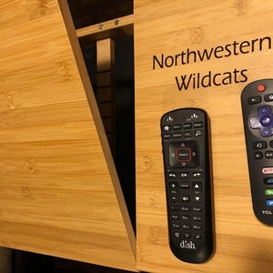 May include: Two black remote controls on a wooden surface. The remote on the left has the word "dish" on it. The remote on the right has the words "Netflix, sling, hulu, vudu, TCL - Roku TV" on it. The surface has the words "Northwestern Wildcats" written on it.