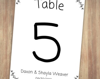 May include: A white table number card with black floral accents and the text "Table 5". The card also includes the names "Daxon & Shayla Weaver" and the date "09/30/2017".