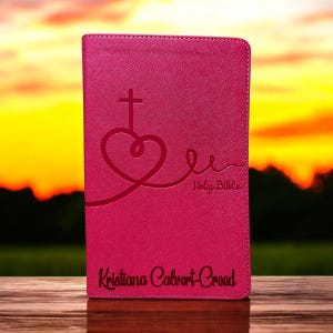 May include: A pink faux leather Holy Bible with a cross and heart design. The name "Kristiana Calvert-Creed" is printed on the front cover.