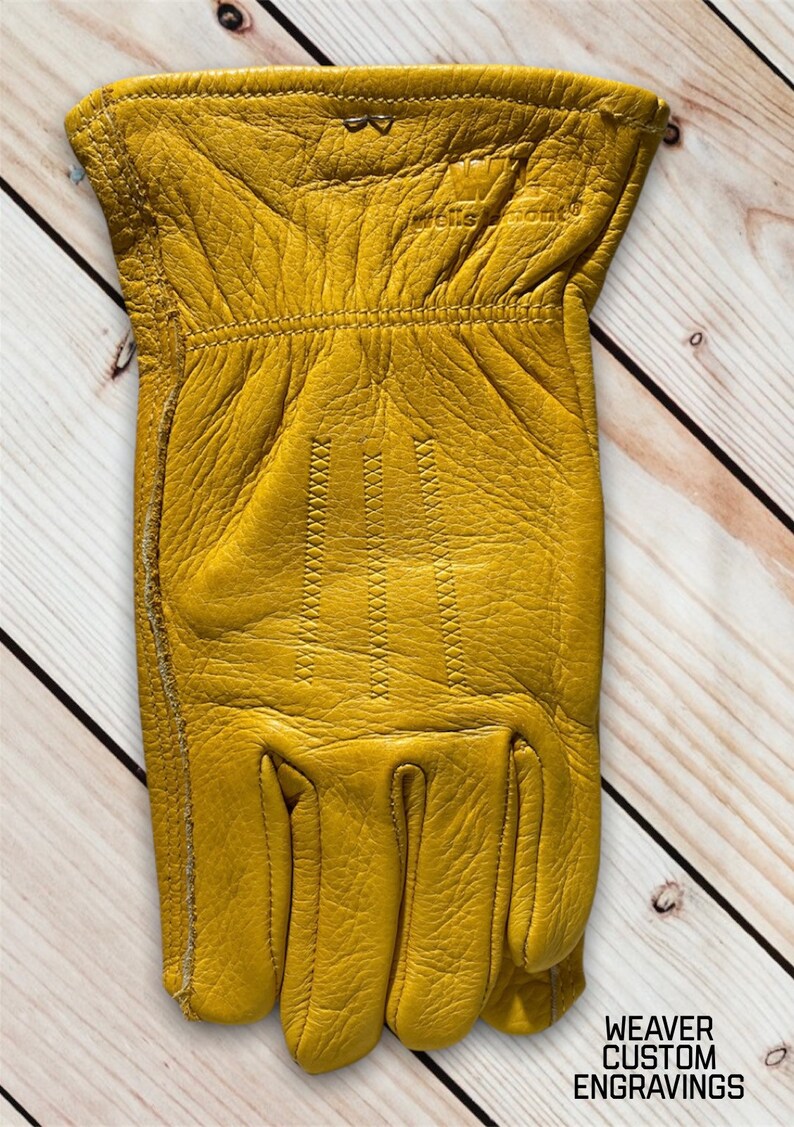 May include: A pair of yellow leather work gloves with stitching detail. The gloves are on a wooden surface.