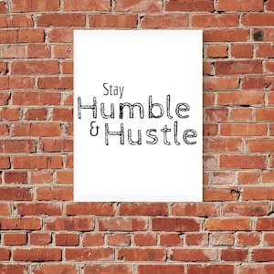 Könnte beinhalten: Weißer gerahmter Druck mit schwarzem Text, der "Stay Humble & Hustle" auf einem Ziegelmauerhintergrund lautet.
