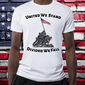 May include: White t-shirt featuring a graphic of soldiers raising the American flag, with the text "UNITED WE STAND DIVIDED WE FALL" in black. The design is a grayscale illustration with a red, white, and blue flag.