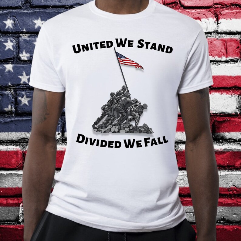 May include: White t-shirt featuring a graphic of soldiers raising the American flag, with the text "UNITED WE STAND DIVIDED WE FALL" in black. The design is a grayscale illustration with a red, white, and blue flag.