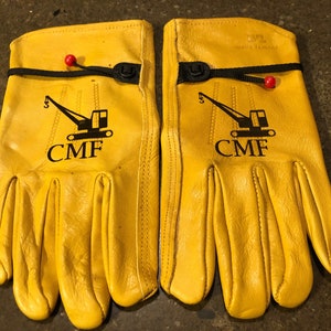 May include: A pair of yellow leather work gloves with black stitching and a black logo that says "CMF" with a crane graphic. The gloves have a black strap with a button closure.