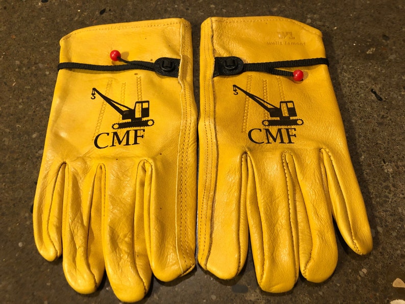 May include: A pair of yellow leather work gloves with black stitching and a black logo that says "CMF" with a crane graphic. The gloves have a black strap with a button closure.