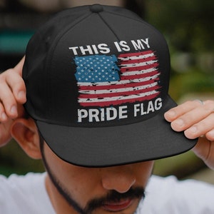 以下が含まれることがあります： 「This is my pride flag」というテキストと、くたびれたアメリカ国旗のグラフィックが描かれた黒いベースボールキャップ。