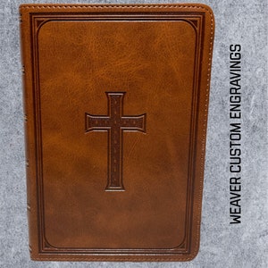 May include: A brown leather-bound journal with a cross embossed on the front cover. The journal has a gold-colored spine and is titled "WEAVER CUSTOM ENGRAVINGS".