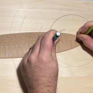 Op de afbeelding: Een persoon gebruikt een potlood en een gereedschap met het opschrift "Easy Circle" om een cirkel te tekenen op een lichtgekleurd houten oppervlak. Het gereedschap is bruin en heeft een reeks gaten langs de rand. Het potlood is groen en wit.