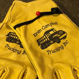 May include: A pair of yellow leather work gloves with a black truck logo and the text "Kevin Campbell Trucking Inc." embroidered on one glove. The other glove has the text "Trucking Inc." embroidered on it.