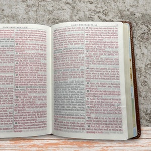 May include: Open brown leather-bound Bible with red text on white pages. The text on the open pages is from the book of Matthew. The Bible is sitting on a wooden surface. The text "WEAVER CUSTOM ENGRAVINGS" is visible on the right side of the image.