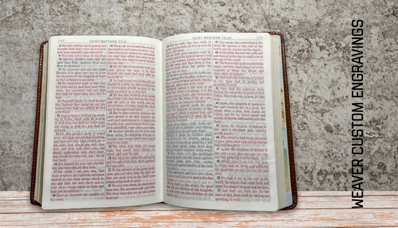 May include: Open brown leather-bound Bible with red text on white pages. The text on the open pages is from the book of Matthew. The Bible is sitting on a wooden surface. The text "WEAVER CUSTOM ENGRAVINGS" is visible on the right side of the image.
