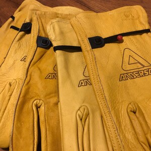 May include: Three pairs of yellow leather work gloves with black adjustable straps and the logo "Anderson" printed on the back of the gloves.