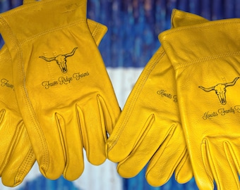 Puede incluir: Dos pares de guantes de trabajo de cuero amarillo con un logotipo de calavera y huesos cruzados. Los guantes están personalizados con el texto "Fawn Ridge Farms" y "Kourtis Family Farms".