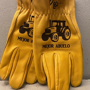May include: A pair of tan leather work gloves with a black tractor design and the text "Mejor Abuelo" on one glove and "Mejor A" on the other glove.