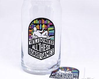 May include: A clear glass can-shaped tumbler featuring a colourful graphic of a person reclining against a bookshelf. The design includes the text "YES, I DO NEED ALL THESE BOARD GAMES" in bold black lettering. A matching sticker is placed in front.