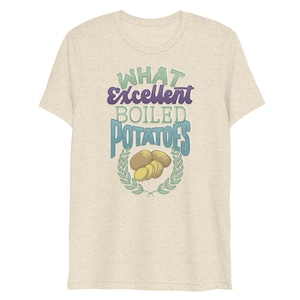 Puede incluir: Una camiseta beige claro con el texto "WHAT Excellent BOILED POTATOES" en azul y morado. El texto está rodeado por una corona de laurel verde con un dibujo de patatas hervidas en el centro.