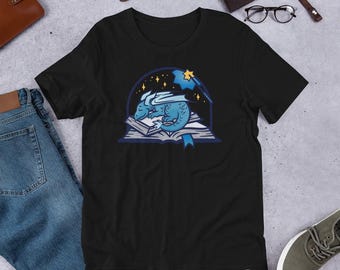 May include: A black short-sleeved t-shirt featuring a graphic print of a small blue dragon sleeping on an open book. The illustration includes a dark blue arched frame, yellow stars, and a yellow magic wand with a blue star tip above the dragon.