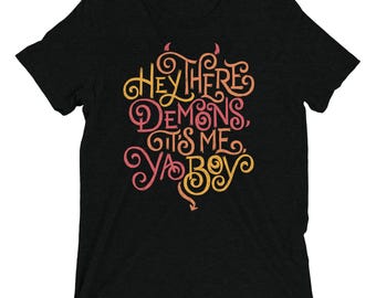 May include: A black short-sleeved t-shirt featuring stylised orange and pink cursive text that reads, Hey There Demons, It's Me, Ya Boy. The lettering includes small decorative devil horns at the top and a pointed tail at the bottom centre.