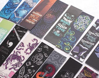 May include: A collection of rectangular paper bookmarks featuring diverse graphic illustrations and typography. Designs include space themes, literary quotes, mythical creatures like Medusa, and abstract patterns in various colours on dark backgrounds.