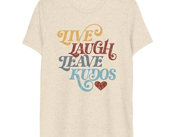 May include: A heathered beige short-sleeve t-shirt featuring the text "LIVE LAUGH LEAVE KUDOS" in stacked, stylised, multi-coloured lettering. The words are printed in yellow, red, and blue, accompanied by a small red heart graphic below the text.