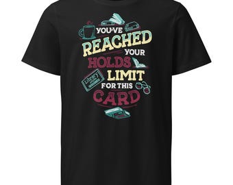 May include: A black short-sleeved t-shirt featuring a graphic design with the text "YOU'VE REACHED YOUR HOLDS LIMIT FOR THIS CARD" in yellow and maroon. The design includes illustrations of a coffee mug, books, a library card, and glasses.