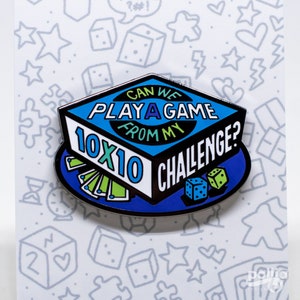 Op de afbeelding: Een blauwe en witte emaille pin met de tekst "Can we play a game from my 10x10 challenge?"  De pin toont een speldoos met dobbelstenen en kaarten.