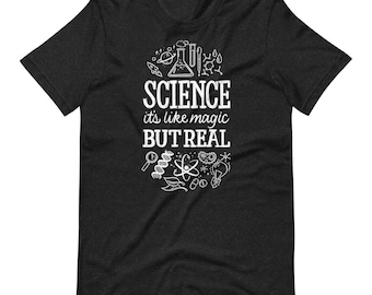 May include: A charcoal grey short-sleeve t-shirt featuring a white graphic print. The design includes the text "SCIENCE it's like magic BUT REAL" surrounded by various scientific illustrations such as a planet, flask, DNA strand, and atom.