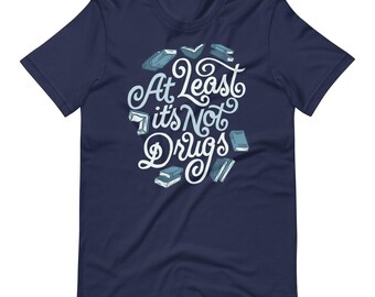 May include: A navy blue short-sleeve t-shirt featuring a graphic print in white and light blue. The design includes the text "At Least it's Not Drugs" in a stylised script font, surrounded by several small illustrations of closed and open books.