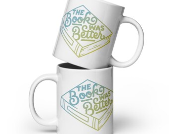 May include: Two white ceramic mugs, one stacked on the other. Each mug displays the text "THE BOOK WAS BETTER" in a blue and yellow gradient design, inside a book-shaped frame. The mugs have a standard handle.
