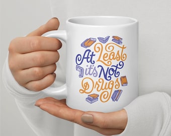 May include: A white ceramic mug featuring the text "At least it's Not Drugs" in a stylised orange and purple font, surrounded by small illustrations of various books in shades of purple and orange scattered around the central lettering.