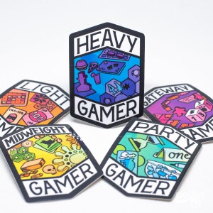 Op de afbeelding: Vijf zeshoekige stickers met verschillende gekleurde achtergronden en illustraties van spelelementen. Elke sticker heeft de tekst "GAMER" en een label dat het type spel beschrijft: "HEAVY", "LIGHT", "MIDWEIGHT", "GATEWAY" en "PARTY".