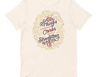 May include: A cream-coloured short-sleeve t-shirt featuring a central graphic of various pasta shapes surrounding the dark red and brown text that reads, I CAN DO ALL THINGS THROUGH CARBS WHICH STRENGTHEN ME. The design is printed on the front chest.