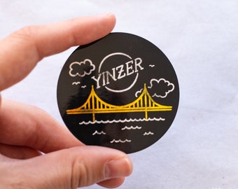 Op de afbeelding: Zwarte en witte sticker met een gele brug, wolken en het woord "YINZER" in een cirkel.